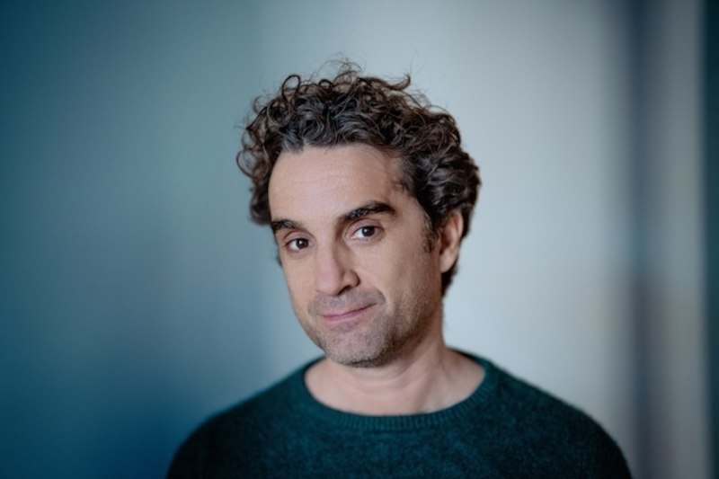 Ein Mann mit lockigem Haar und grünem Pullover blickt leicht lächelnd in die Kamera. Der Hintergrund ist unscharf und blau-grau., © Renitenztheater Stuttgart e.V.