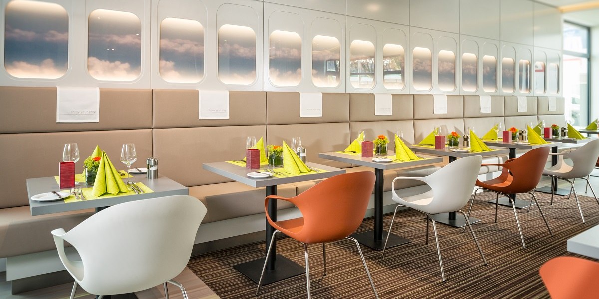 Modern restaurant with airplane window decor, tables set with green napkins and colorful chairs., &copy; Essential by Dorint