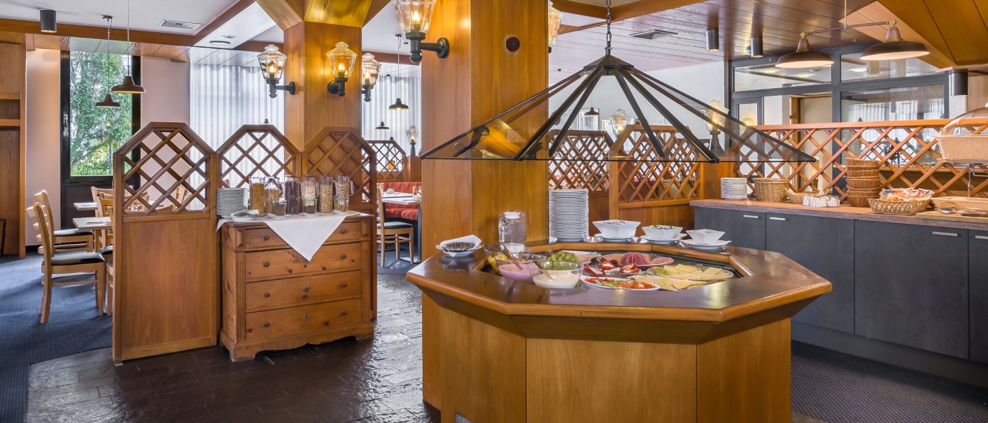 Ein Frühstücksbuffet in einem holzgetäfelten Raum mit Tischen, Stühlen und verschiedenen Speisen auf einem zentralen Tisch., © Novum Management GmbH Ein Frühstücksbuffet in einem holzgetäfelten Raum mit Tischen, Stühlen und verschiedenen Speisen auf einem zentralen Tisch., © Novum Management GmbH