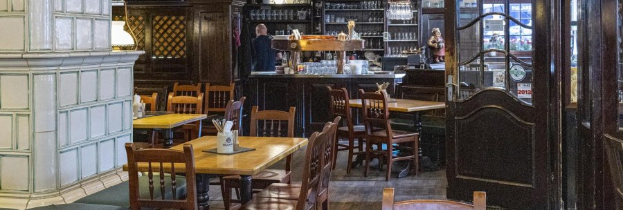 Gemütlicher Innenraum eines Restaurants mit Holztischen und -stühlen, einer Bar im Hintergrund und einem großen Kachelofen links., © SMG, Sarah Schmid