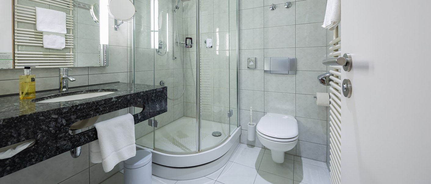 Modernes Badezimmer mit Glasdusche, Waschbecken mit dunkler Ablage, Toilette und Handtuchhalter. Helle Fliesen und stilvolle Armaturen., © Kronenhotel Stuttgart