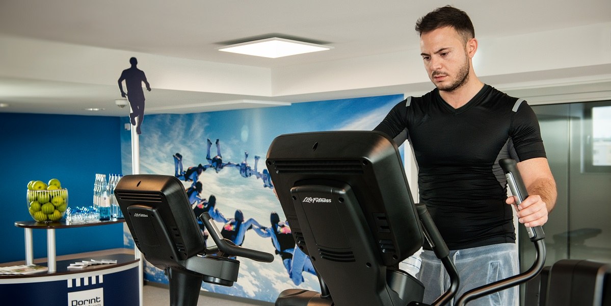 A man trains on a cross trainer in a fitness room. In the background is a blue wall with a parachutist motif., &copy; Essential by Dorint
