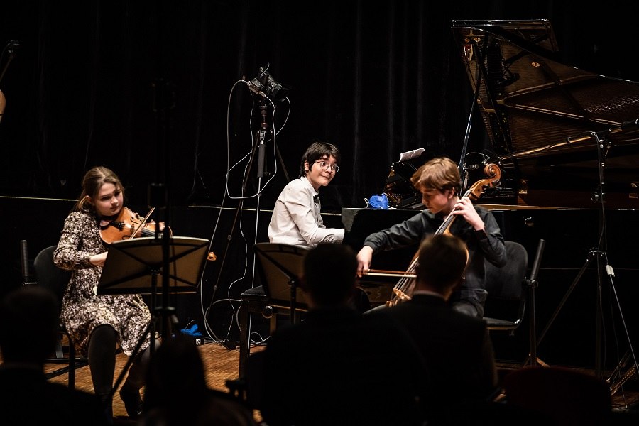 Drei junge Musiker spielen Violine, Klavier und Cello auf einer Bühne vor Publikum. Im Hintergrund steht ein Flügel., © unbekannt