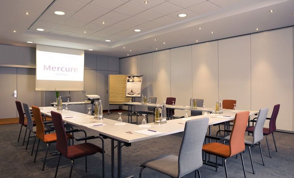 Modern conference room with U-shaped table arrangement, chairs in different colors and presentation technology. A projector image with 'Mercure Hotels' on the wall., &copy; Mercure Stuttgart Airport Messe