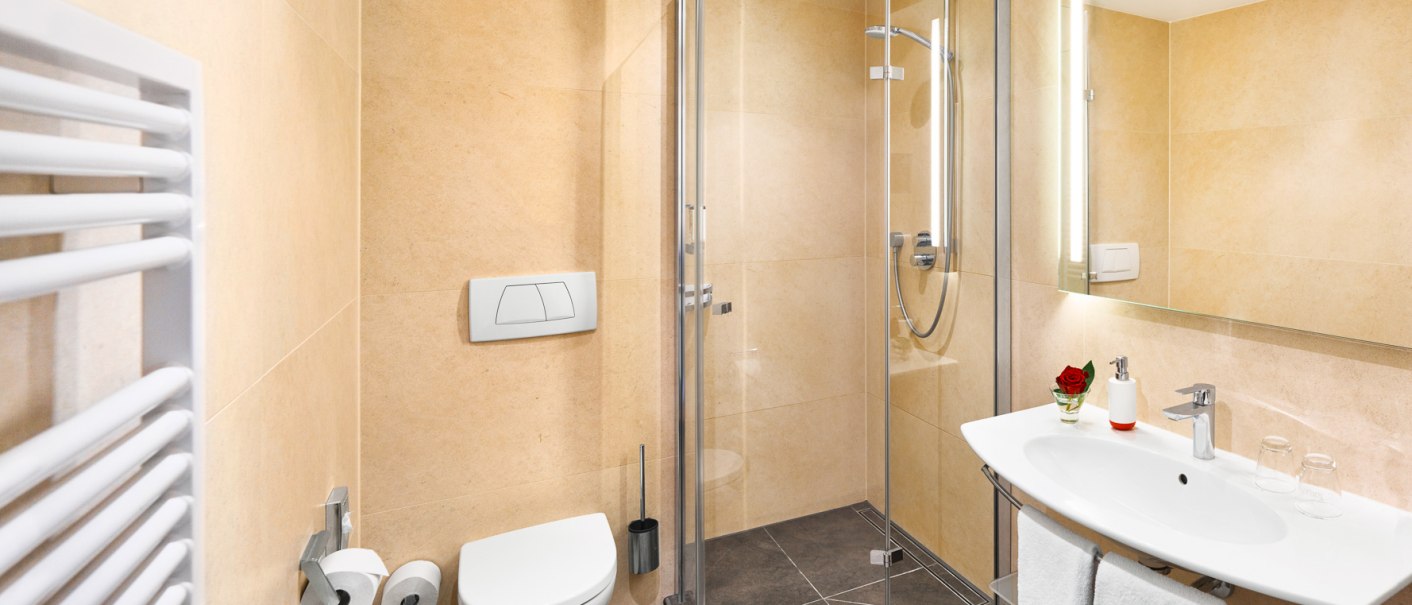 Modernes Badezimmer mit Glasdusche, Waschbecken, WC und Handtuchhalter. Beige Fliesen, Spiegel mit Beleuchtung und rote Rose als Dekoration., © TOMAS