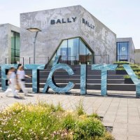 Large letters spell out the word 'OUTLETCITY' in front of modern buildings. People walk by while flowers bloom in the foreground., &copy; OUTLETCITY AG