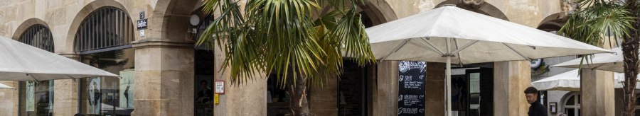 Café with tables and chairs under parasols in front of a historic building with arcades. Palm trees and people in the outdoor area., © SMG, Sarah Schmid
