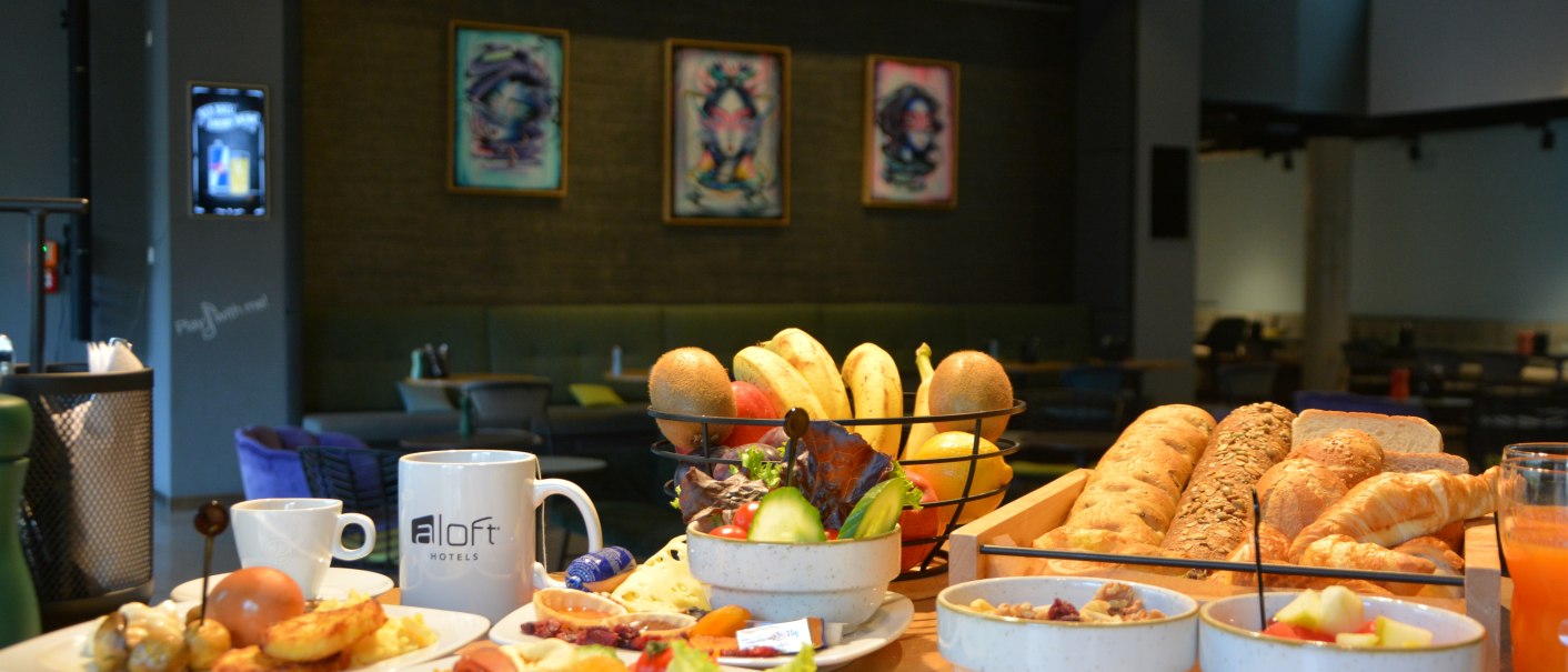 Ein üppiges Frühstücksbuffet in einem Hotelrestaurant mit Brot, Obst, Müsli und Getränken. Im Hintergrund sind moderne Kunstwerke zu sehen., © Aloft Stuttgart Ein üppiges Frühstücksbuffet in einem Hotelrestaurant mit Brot, Obst, Müsli und Getränken. Im Hintergrund sind moderne Kunstwerke zu sehen., © Aloft Stuttgart