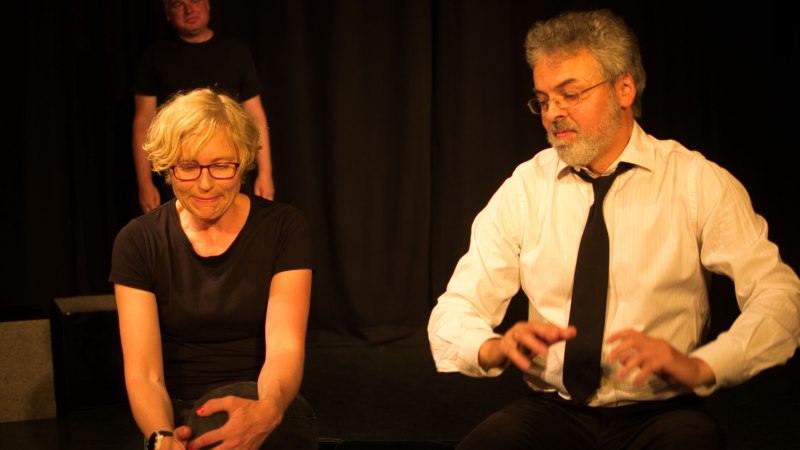 Drei Schauspieler auf einer Bühne, zwei sitzen im Vordergrund, eine Person steht im Hintergrund. Die Szene wirkt intensiv und konzentriert., © Die Maßschneiderei Drei Schauspieler auf einer Bühne, zwei sitzen im Vordergrund, eine Person steht im Hintergrund. Die Szene wirkt intensiv und konzentriert., © Die Maßschneiderei