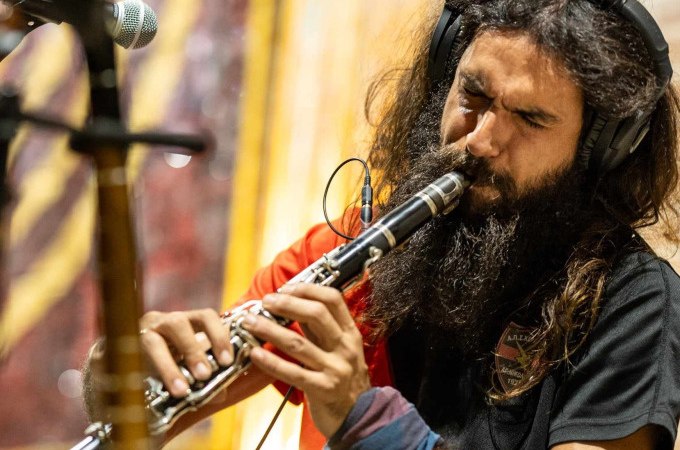 A musician with long hair and a beard is concentrating on playing the clarinet. He is wearing headphones and a black T-shirt. A microphone can be seen in the background., &copy; Landesmuseum W&uuml;rttemberg