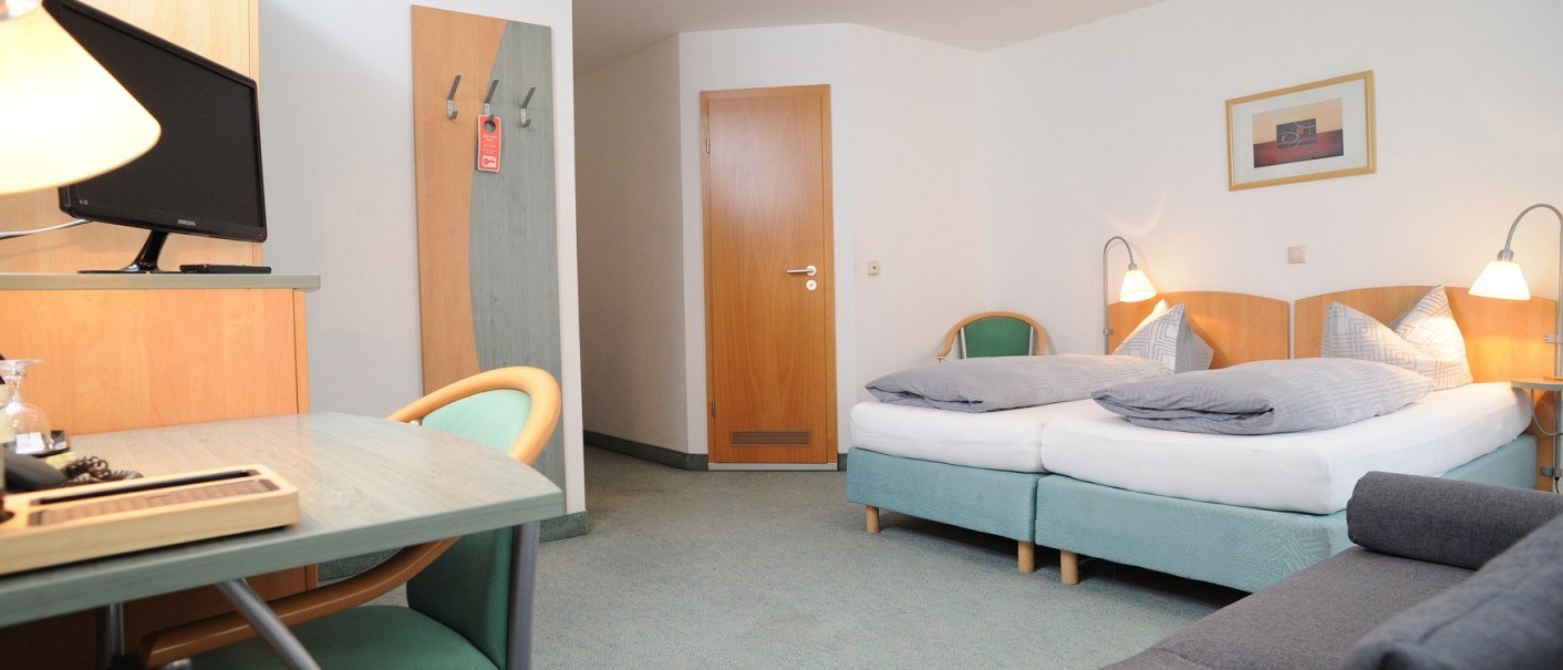 A hotel room with double bed, gray sofa, desk with TV and modern furnishings. Light-colored walls and carpeted floor., © TOMAS