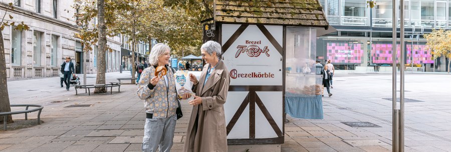 Two women are laughing in front of a pretzel stand on Schlossplatz. One is holding a pretzel, the other a bag. Autumnal trees in the background., &copy; SMG, Sarah Schmid