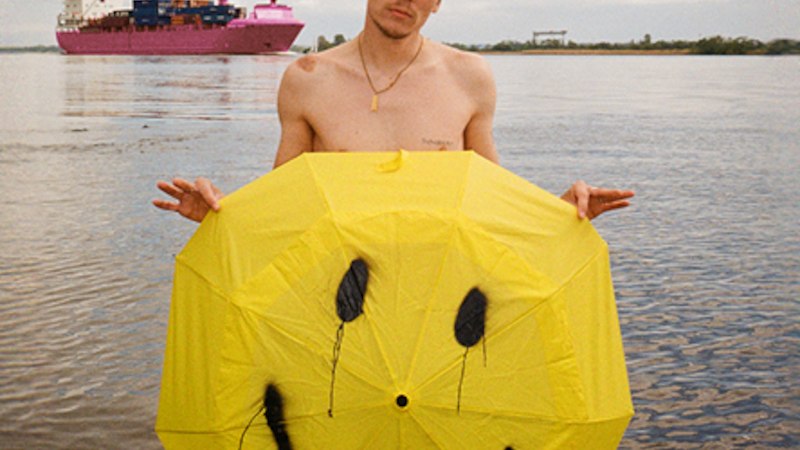 A man with sunglasses stands in the water holding a yellow umbrella with a smiley face. A large ship passes by in the background., © Rosenau Kultur e.V. A man with sunglasses stands in the water holding a yellow umbrella with a smiley face. A large ship passes by in the background., © Rosenau Kultur e.V.