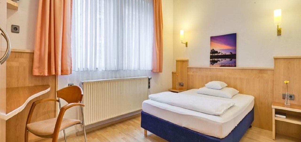 Ein gemütliches Hotelzimmer mit Einzelbett, Holzmöbeln, Stuhl und Bild an der Wand. Große Fenster mit Vorhängen lassen Licht herein., © ARNOTEL GmbH