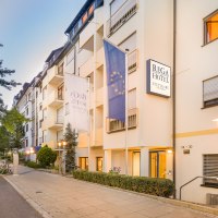 Außenansicht des Rega Hotels in Stuttgart. Mehrstöckiges Gebäude mit Fahnen und beleuchteten Straßenlaternen. Ruhige Straße mit geparkten Autos., © TOMAS Außenansicht des Rega Hotels in Stuttgart. Mehrstöckiges Gebäude mit Fahnen und beleuchteten Straßenlaternen. Ruhige Straße mit geparkten Autos., © TOMAS