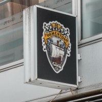 A sign with the Cape Tormentoso logo hangs on a modern building. It shows a sailing ship and decorative elements., © Stuttgart Marketing GmbH, Sarah Schmid A sign with the Cape Tormentoso logo hangs on a modern building. It shows a sailing ship and decorative elements., © Stuttgart Marketing GmbH, Sarah Schmid