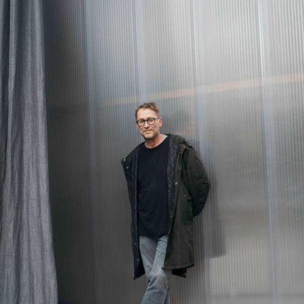 Ein Mann mit Brille und dunklem Mantel lehnt lässig an einer strukturierten Metallwand. Er trägt ein schwarzes Shirt und graue Hose., © Theaterhaus Stuttgart e.V. Ein Mann mit Brille und dunklem Mantel lehnt lässig an einer strukturierten Metallwand. Er trägt ein schwarzes Shirt und graue Hose., © Theaterhaus Stuttgart e.V.