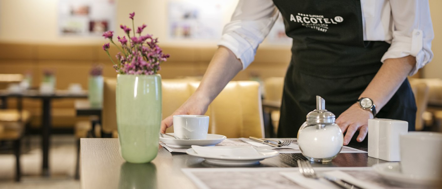 Eine Person deckt einen Tisch in einem Restaurant. Auf dem Tisch stehen eine Tasse, eine Zuckerdose und eine Vase mit Blumen., © Arcotel Hotels Eine Person deckt einen Tisch in einem Restaurant. Auf dem Tisch stehen eine Tasse, eine Zuckerdose und eine Vase mit Blumen., © Arcotel Hotels