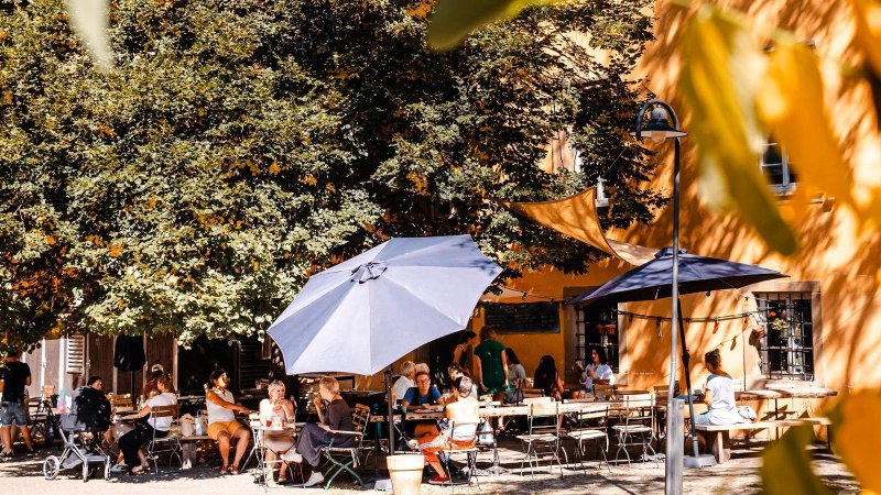 Menschen genießen das sonnige Wetter in einem Café im Freien, umgeben von Bäumen und Sonnenschirmen. Eine entspannte Atmosphäre herrscht vor., © Stuttgart-Marketing GmbH, Sarah Schmid