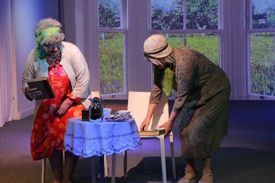 Two women in costumes stand on a stage. One is holding a book, the other is straightening a chair. There is a coffee pot and cups on the table., &copy; Foto_Katja Ritter