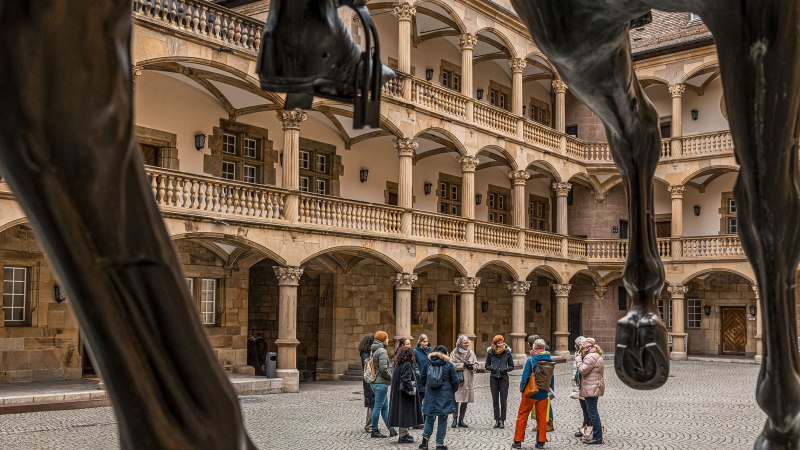 Innenhof eines alten Schlosses mit Arkaden und Säulen. Eine Gruppe von Menschen steht zusammen, im Vordergrund sind die Beine einer Pferdestatue sichtbar., © Stuttgart Marketing GmbH - Sarah Schmid Innenhof eines alten Schlosses mit Arkaden und Säulen. Eine Gruppe von Menschen steht zusammen, im Vordergrund sind die Beine einer Pferdestatue sichtbar., © Stuttgart Marketing GmbH - Sarah Schmid