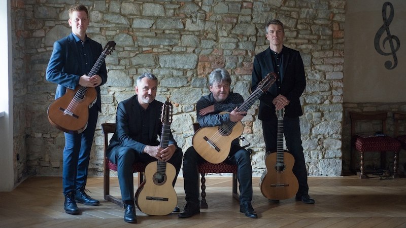 Vier Männer mit Gitarren posieren vor einer Steinwand in einem Raum mit Holzboden. Zwei sitzen, zwei stehen, alle tragen dunkle Kleidung., © Staatliche Hochschule für Musik und Darstellende Kunst Vier Männer mit Gitarren posieren vor einer Steinwand in einem Raum mit Holzboden. Zwei sitzen, zwei stehen, alle tragen dunkle Kleidung., © Staatliche Hochschule für Musik und Darstellende Kunst