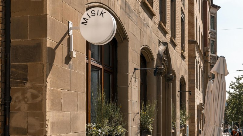 Geb&auml;ude mit Sandsteinfassade, rundes Schild mit 'MISIK', Fenster mit Pflanzen, geschlossene Sonnenschirme auf der Stra&szlig;e., &copy; Stuttgart-Marketing GmbH, Sarah Schmid