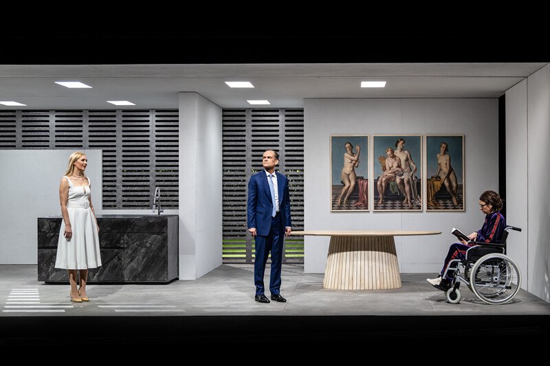 Theater scene in a modern room with three people. A woman in a wheelchair reads while a man and a woman stand. Works of art on the wall., &copy; W&uuml;rttembergische Staatstheater Stuttgart