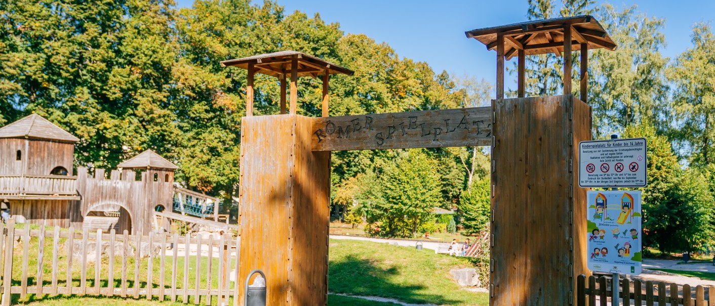 Eingang zum Römer-Spielplatz mit Holztor und Schildern. Im Hintergrund sind Bäume und ein hölzernes Spielgerüst zu sehen., © Stuttgart-Marketing GmbH, Thomas Niedermüller