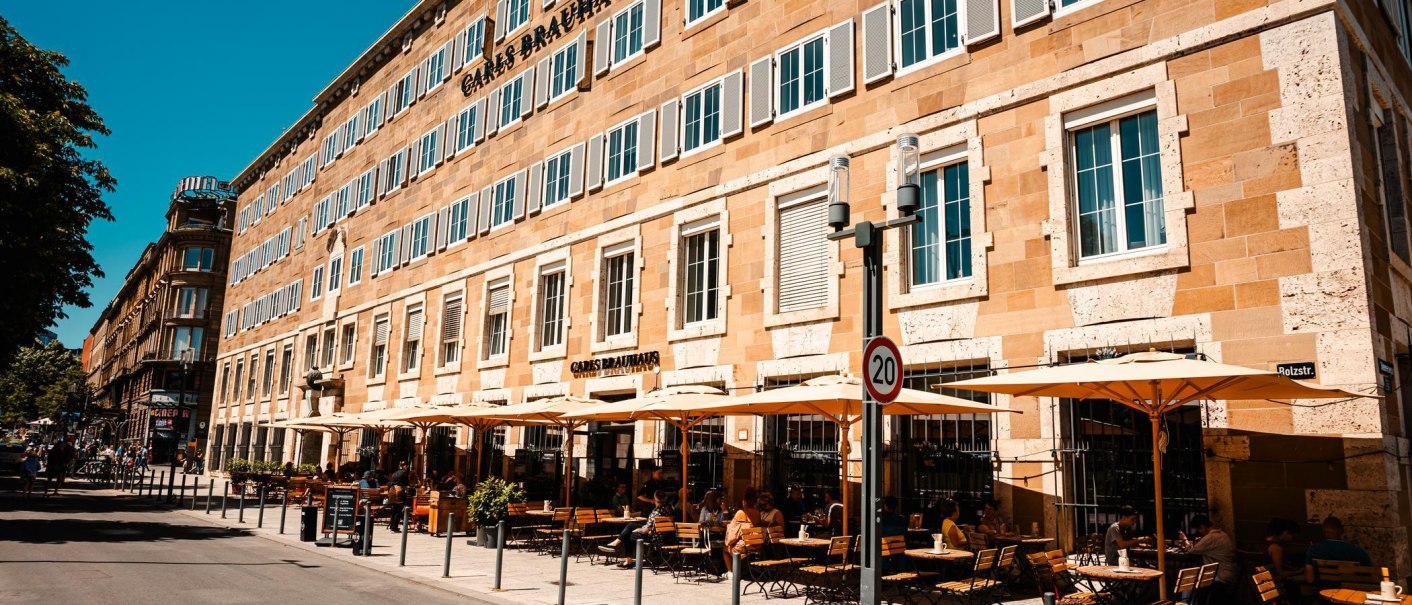 Carls Brauhaus mit gelbem Gebäude, vielen Fenstern und Außenterrasse mit Sonnenschirmen und Sitzplätzen. Menschen genießen das sonnige Wetter., © Stuttgart Marketing GmbH, Sarah Schmid