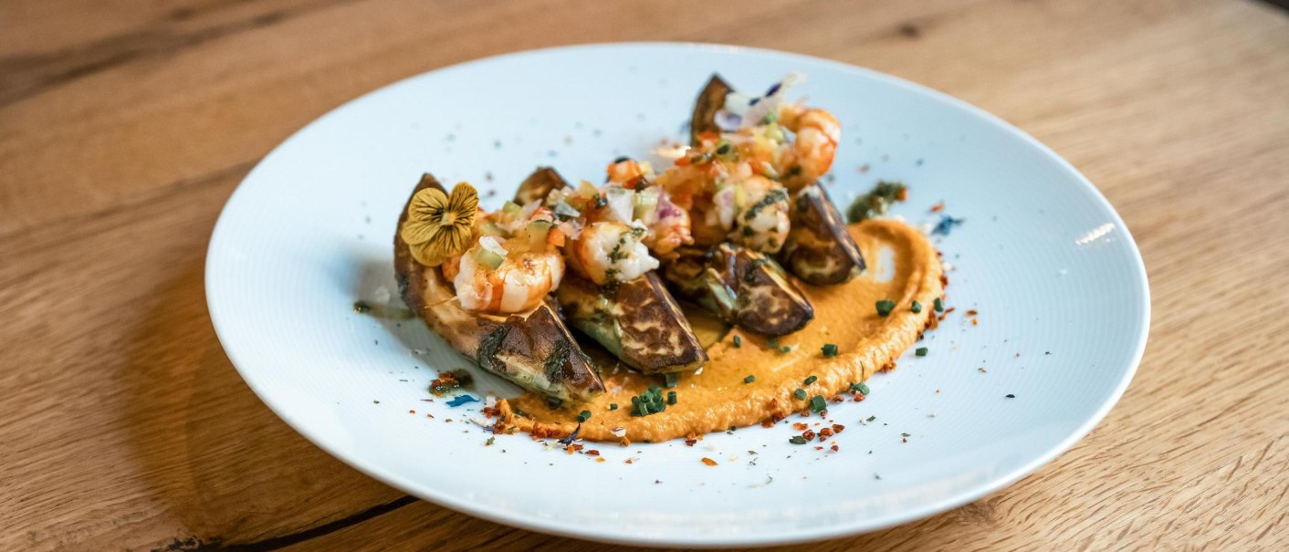 Ein Teller mit kunstvoll angerichteten Tapas, darunter Garnelen und Gemüse auf einer cremigen Sauce, dekoriert mit essbaren Blüten, auf einem Holztisch., © Jose y Josefina, Stuttgart