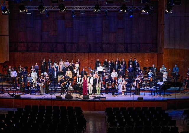 Ein Orchester mit Chor steht auf einer Bühne in einem Konzertsaal. Die Musiker tragen festliche Kleidung, der Saal ist mit leeren Stuhlreihen gefüllt., © Marc Schäfer