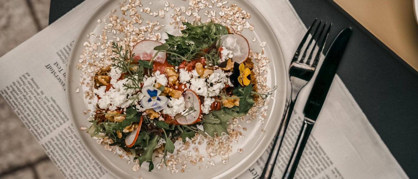 Ein kunstvoll angerichtetes Frühstück mit Salat, Radieschen, Feta und essbaren Blumen auf einem Teller, umgeben von Haferflocken, auf einer Zeitung serviert., © Fritz Stuttgart