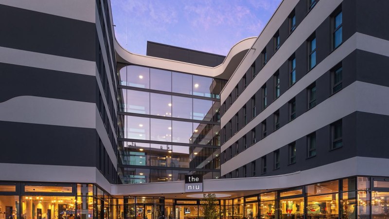 A modern hotel exterior with a glass facade and an illuminated entrance area at dusk. The building features an elegant, wave-like design., © Novum Hospitality A modern hotel exterior with a glass facade and an illuminated entrance area at dusk. The building features an elegant, wave-like design., © Novum Hospitality
