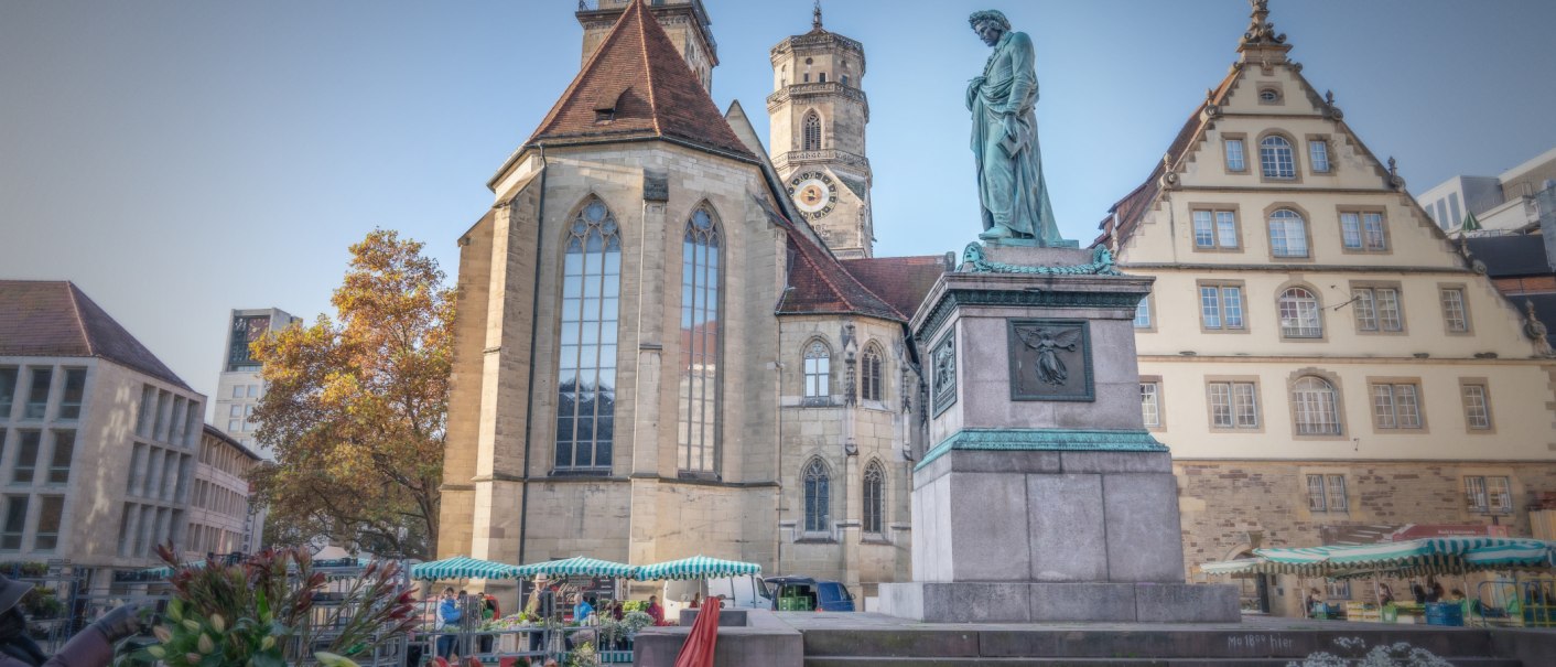 Der Schillerplatz in Stuttgart zeigt eine Statue, historische Gebäude und Marktstände. Im Hintergrund ist eine Kirche mit Turm zu sehen., © SMG Martina Denker Der Schillerplatz in Stuttgart zeigt eine Statue, historische Gebäude und Marktstände. Im Hintergrund ist eine Kirche mit Turm zu sehen., © SMG Martina Denker