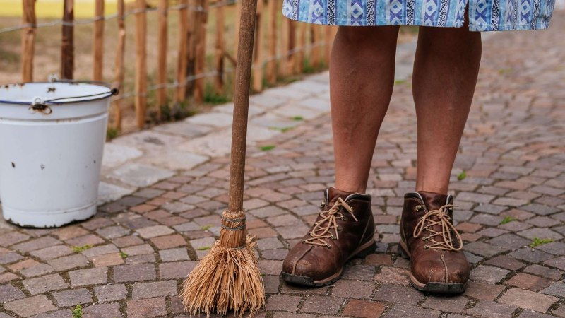 Person in braunen Schuhen steht auf Kopfsteinpflaster, hält einen Besen. Ein weißer Eimer steht daneben. Die Person trägt einen blauen Rock., © Thomas Niedermüller