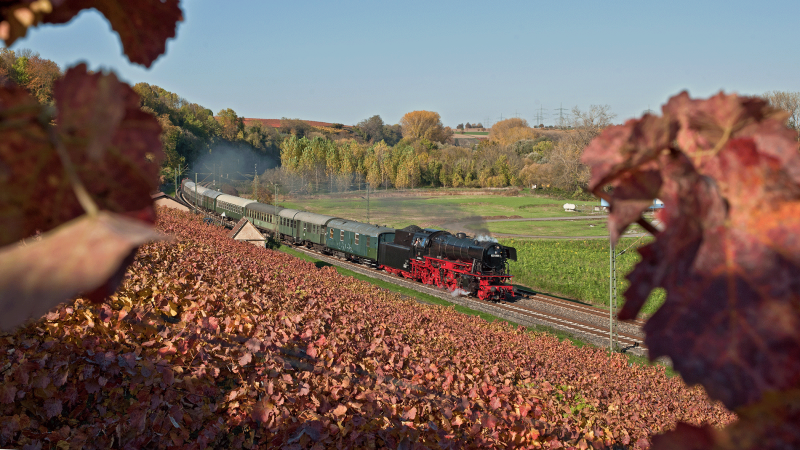 A steam locomotive pulls a train through an autumnal landscape with colorful vineyards and trees., © DBK Historische Bahn e.V. A steam locomotive pulls a train through an autumnal landscape with colorful vineyards and trees., © DBK Historische Bahn e.V.