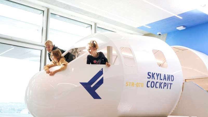 Drei Kinder spielen in einem Cockpit-Nachbau mit der Aufschrift 'SkyLand Cockpit' und blicken aus den Fenstern. Helle, moderne Umgebung., © Flughafen Stuttgart