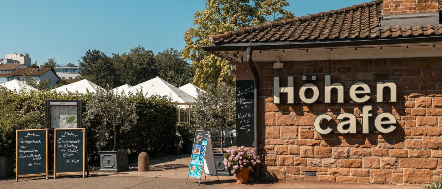 Das Höhencafé mit Speisekarten und Sonnenschirmen im Hintergrund. Die Fassade ist aus Backstein, und es gibt Angebote für Pommes und Eis., © Stuttgart Marketing GmbH, Sarah Schmid