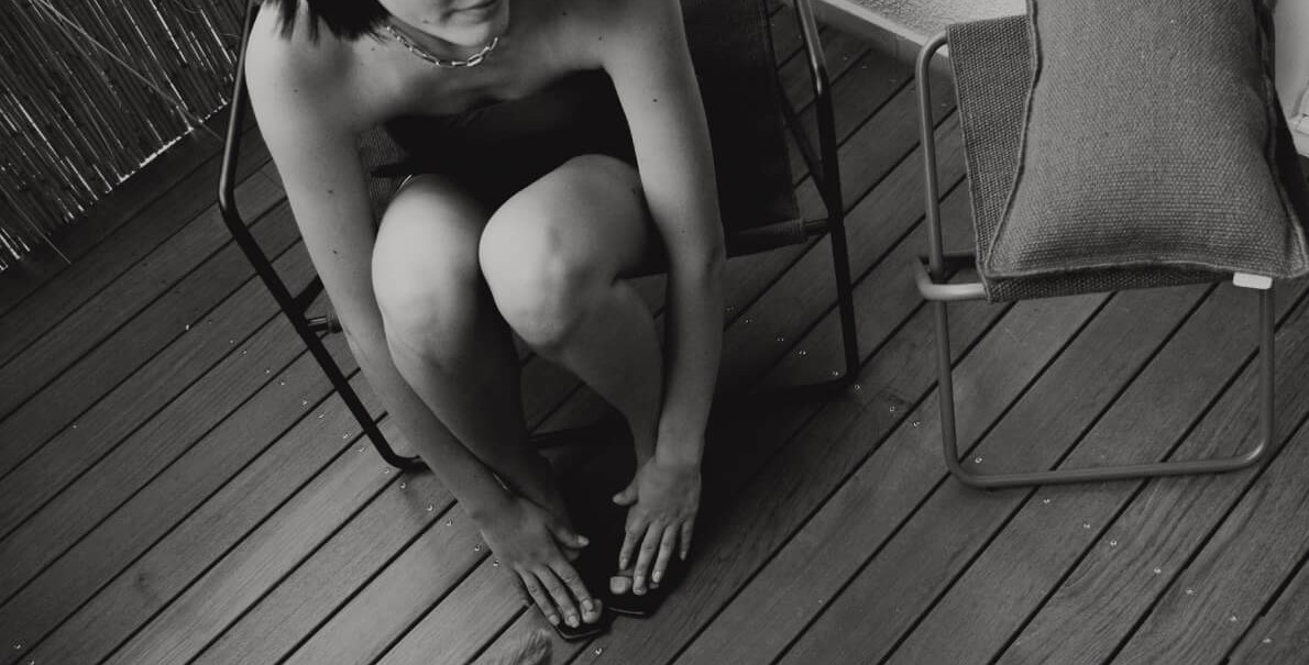 Eine Person sitzt auf einem Balkonstuhl und beugt sich zu einem Hund hinunter. Der Balkon hat Holzboden und eine Bambuswand. Schwarz-wei&szlig;-Foto., &copy; Peter Oliver Wolff / DAS EDITH