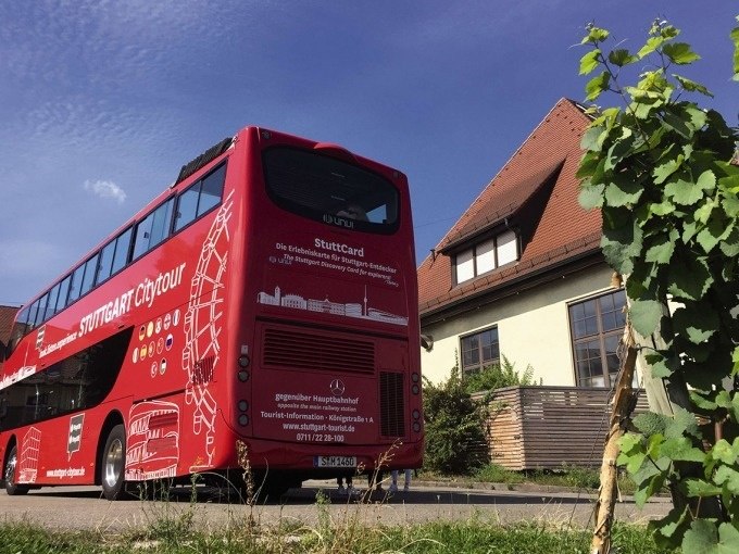 Roter Doppeldeckerbus der Stuttgart Citytour vor einem Haus mit rotem Dach und blauen Himmel im Hintergrund., © Willms Touristik Roter Doppeldeckerbus der Stuttgart Citytour vor einem Haus mit rotem Dach und blauen Himmel im Hintergrund., © Willms Touristik