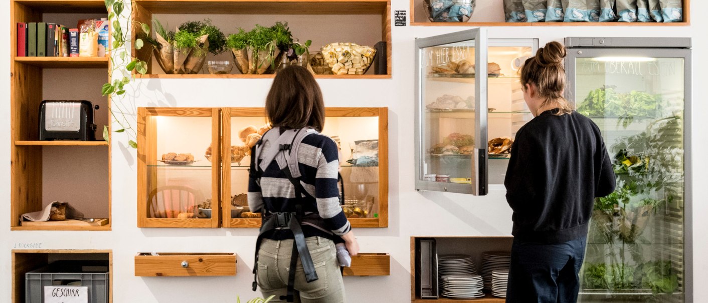 Two people are looking at shelves with food and plants. A refrigerator and dishes are also visible., © Sven Weber