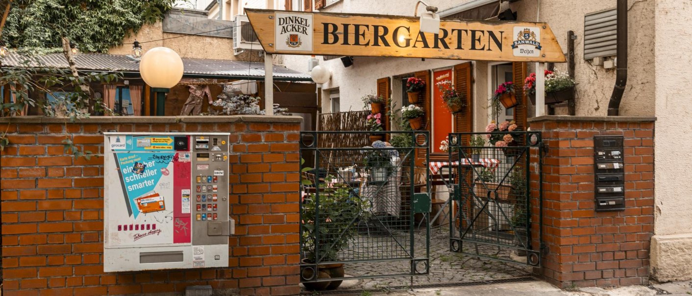 Eingang zu einem Biergarten mit Ziegelmauer, Zigarettenautomat und Blumen. Ein Schild mit "Biergarten" hängt über dem Tor., © SMG, Sarah Schmid