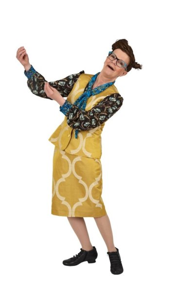 Woman in yellow outfit with blue glasses and scarf poses cheerfully. She is wearing a patterned top and black shoes., &copy; Rosenau Kultur e.V.