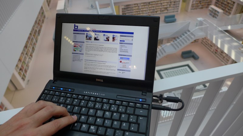 Ein Laptop zeigt eine Webseite in einer modernen Bibliothek. Im Hintergrund sind weiße Regale und Treppen zu sehen., © Stadtbibliothek Stuttgart