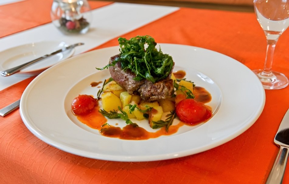 An elegant dish of meat, potatoes, tomatoes and green salad on a white plate, served on an orange table., © Hotel Neuwirtshaus An elegant dish of meat, potatoes, tomatoes and green salad on a white plate, served on an orange table., © Hotel Neuwirtshaus