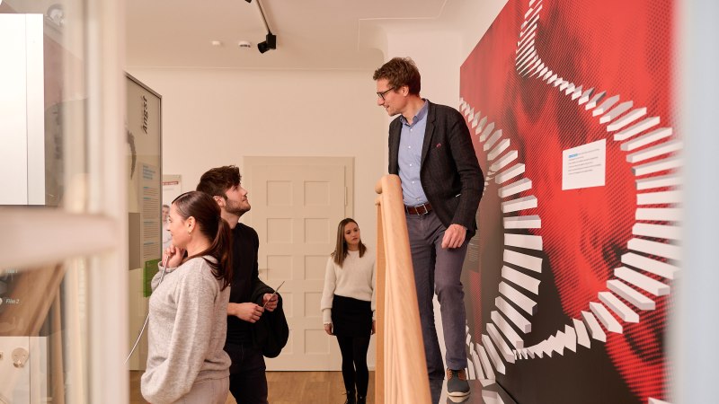 Menschen in einem Ausstellungsraum. Eine Person steht auf einer Treppe, andere betrachten eine Wand mit rotem Design. Thema: Angst., © Stiftung Psyche Menschen in einem Ausstellungsraum. Eine Person steht auf einer Treppe, andere betrachten eine Wand mit rotem Design. Thema: Angst., © Stiftung Psyche