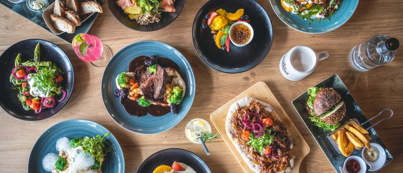 Ein Tisch mit verschiedenen Gerichten: Fleisch mit Gem&uuml;se, Salate, Burger mit Pommes, Brot und Desserts. Getr&auml;nke sind ebenfalls zu sehen., &copy; SatzDrei