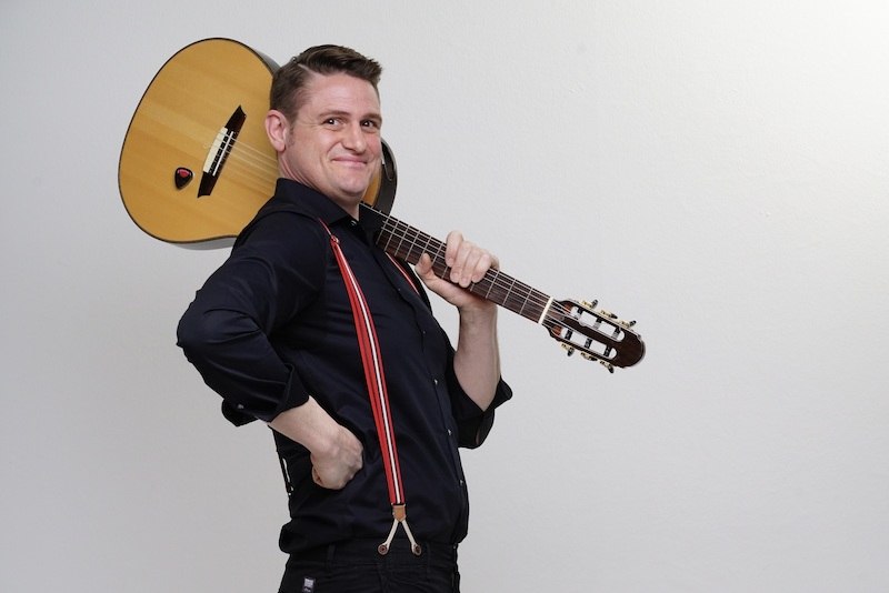 Ein Mann trägt eine Gitarre auf der Schulter und lächelt. Er trägt ein schwarzes Hemd und rote Hosenträger. Der Hintergrund ist schlicht weiß., © Theaterhaus Stuttgart e.V.