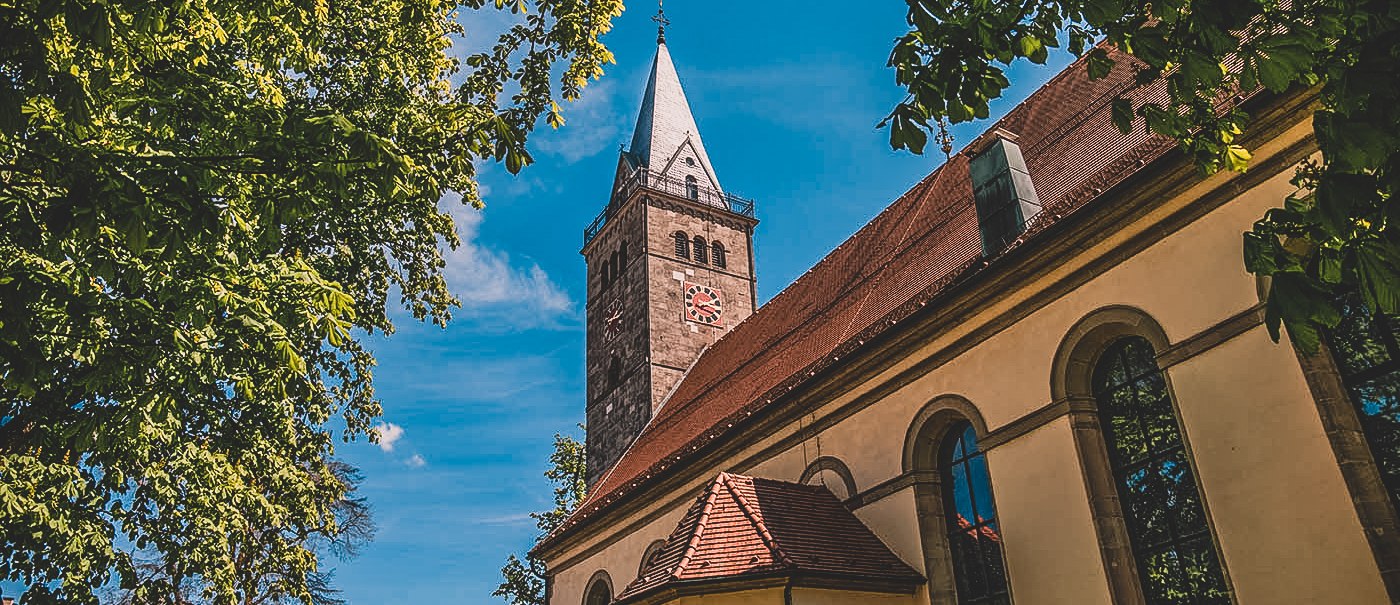 A church tower rises into the blue sky, surrounded by green trees. A historic building with large windows can be seen in the foreground., &copy; Stadt Welzheim
